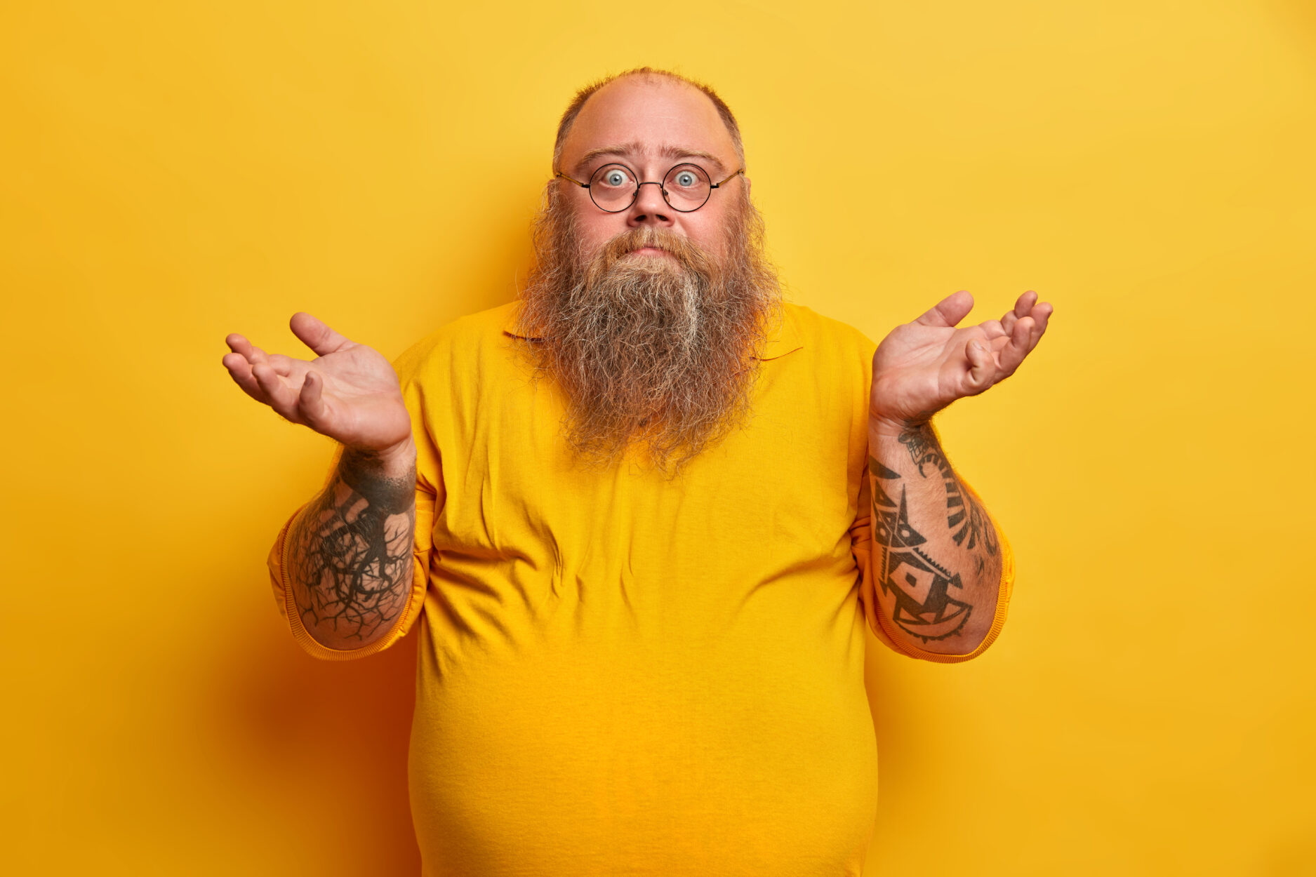 Indoor shot of hesitant bearded man with excess weight shrugs shoulders and stands unaware, has thick beard, big beer belly, dressed in yellow t shirt, round spectacles, faces difficult choice.